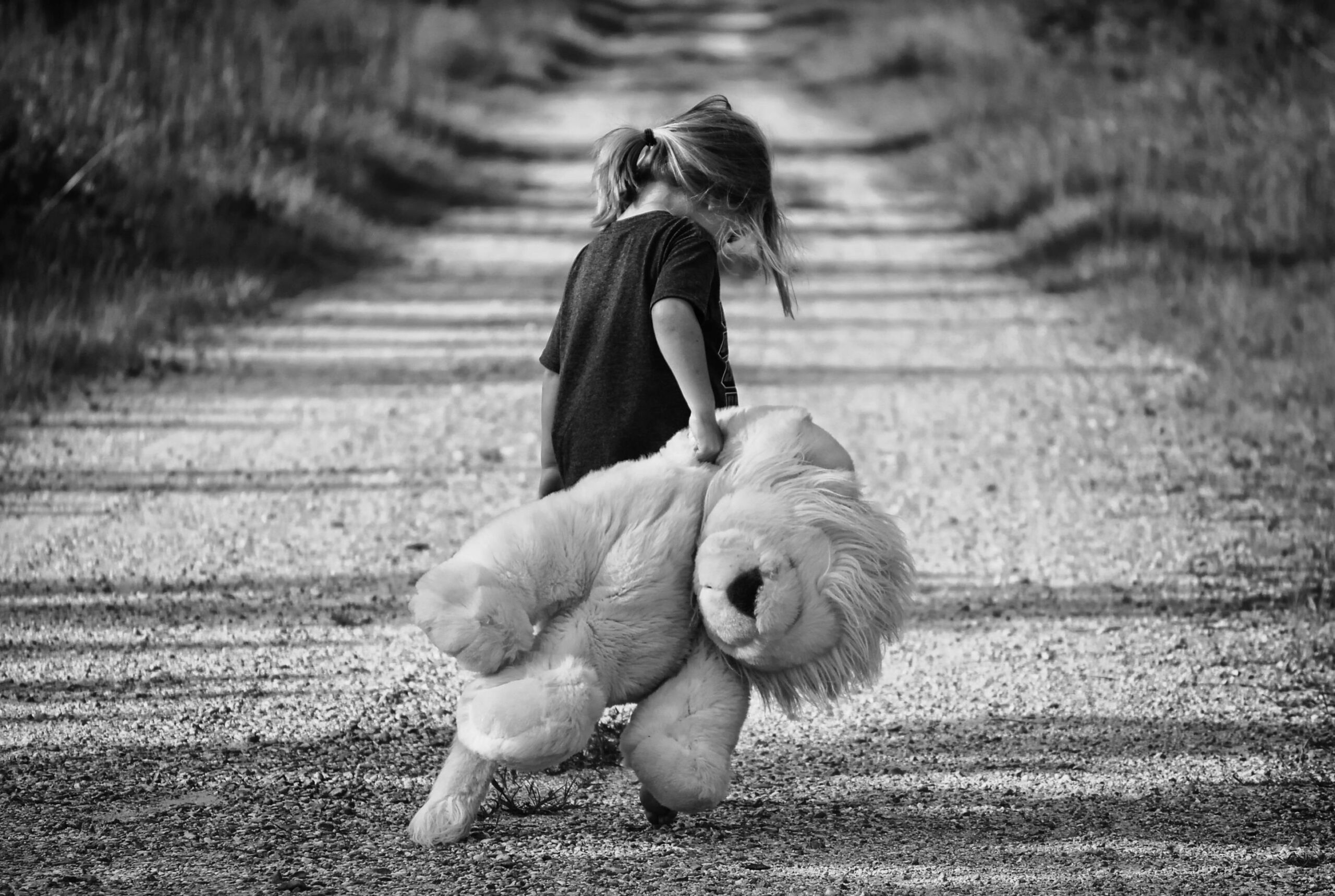 Monochrome image of a child holding a teddy bear while walking down a dirt road, conveying a sense of solitude.